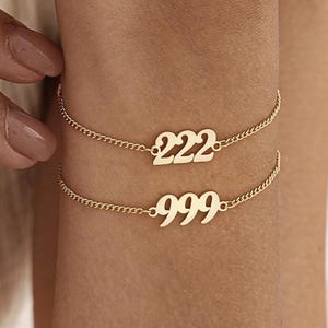 May include: Two gold-colored chain bracelets. The top bracelet displays the number "222", and the bottom bracelet displays the number "999". The bracelets are worn on a wrist.