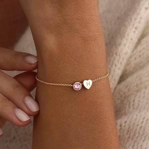 Op de afbeelding: Een delicate gouden kettingarmband met een kleine, ronde, roze edelsteen in een zetting naast een piepkleine, gepolijste gouden hartvormige bedel gegraveerd met de letter H. Het armbandje bestaat uit een fijne schakelketting op de huid.