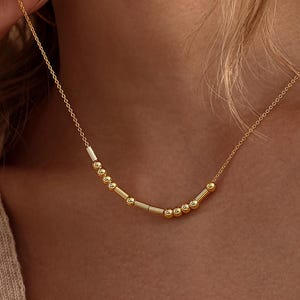 May include: A gold-toned necklace featuring a delicate chain with a central segment composed of small round beads and short cylindrical bars arranged in a sequence, resting against the neck with a subtle metallic finish and a refined appearance.