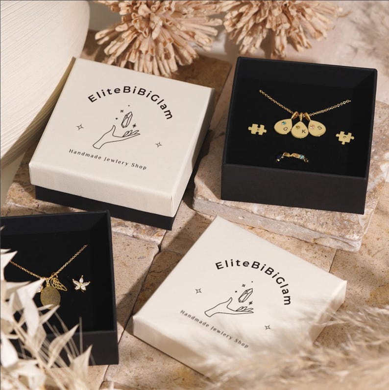 May include: Two square jewelry boxes with cream lids featuring the text EliteBiBiGlam and a hand illustration. One box contains a gold necklace with three teardrop charms, two puzzle piece earrings, and a ring. Another box holds a gold necklace.