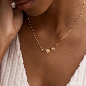 May include: A gold-toned necklace featuring a delicate chain and three small, open-work flower charms. The center flower is slightly larger than the two flanking it, and each flower contains a tiny, clear, sparkling stone at its center.