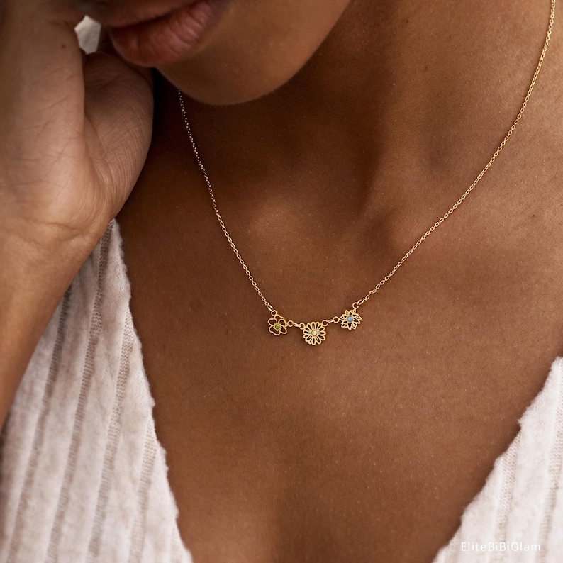 May include: A gold-toned necklace featuring a delicate chain and three small, open-work flower charms. The center flower is slightly larger than the two flanking it, and each flower contains a tiny, clear, sparkling stone at its center.