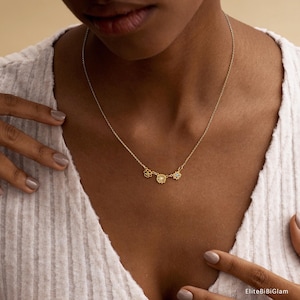 May include: A delicate gold-tone necklace featuring three small, distinct flower-shaped charms centered along a thin chain. The necklace rests against a white ribbed top. The text EliteBiBiGlam is visible in the bottom right corner of the image.