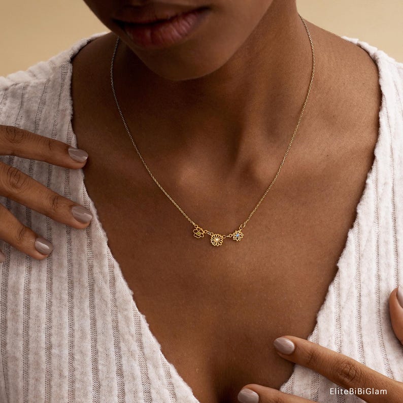 May include: A delicate gold-tone necklace featuring three small, distinct flower-shaped charms centered along a thin chain. The necklace rests against a white ribbed top. The text EliteBiBiGlam is visible in the bottom right corner of the image.