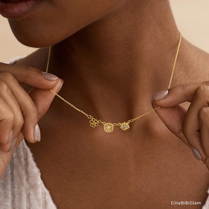 May include: A gold-toned chain necklace featuring three small, distinct flower-shaped charms. The center charm is a daisy design with a small white stone, flanked by two other floral motifs, one with a yellow stone and one with a blue stone.