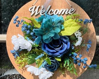 Può includere: Un cartello di benvenuto rotondo in legno con un arrangiamento floreale blu, bianco e verde. Il cartello ha la scritta "Welcome" in lettere bianche.