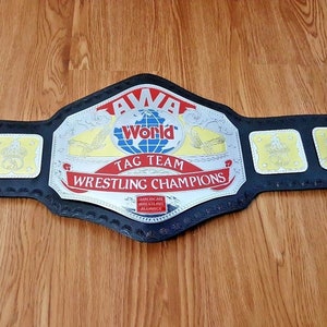 May include: AWA World Tag Team Wrestling Champions belt with a silver and black design. The belt has a large central plate with the words "AWA World Tag Team Wrestling Champions" and a smaller plate with the words "AWA World". The belt has four smaller plates with wrestlers on them.