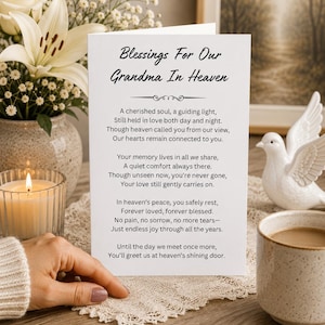 May include: A white folded sympathy card featuring the black printed text Blessings For Our Grandma In Heaven followed by a memorial poem. The card is displayed upright on a lace doily, surrounded by a lit candle, white flowers, and a white dove figurine.