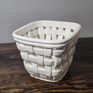 Peut inclure: Un panier carré en céramique blanche avec un motif en treillis tressé. Le récipient présente une finition lisse et brillante ainsi qu'un bord supérieur épais et solide, avec une structure ajourée formant un motif en grille sur les côtés.