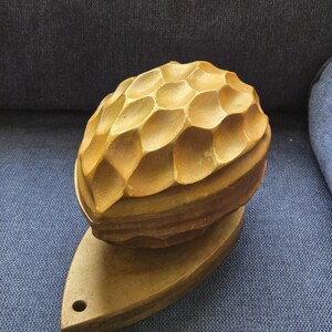 May include: A wooden nutcracker with a textured, patterned top and a flat, boat-shaped base. The nutcracker is a warm, golden brown color and appears to be hand-carved. The base has a small hole near the pointed end.