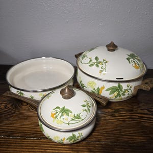 May include: A set of vintage enamelware cookware. The set includes a skillet, a saucepan with a lid, and a pot with a lid. Each piece is white with a floral design in green and yellow, and bronze-colored handles.