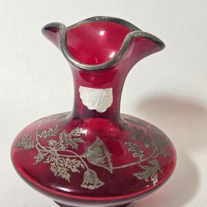 May include: A small ruby red glass vase with a ruffled rim and a silver metallic overlay featuring a floral pattern. The vase has a narrow neck and a wide, rounded base. A small, partially obscured white rectangular label is attached to the neck.