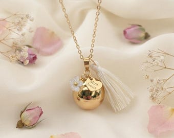 Könnte beinhalten: Eine goldfarbene Halskette mit einem kugelförmigen Anhänger, einer zarten Kette, einem kleinen weißen Blumenanhänger, einem herzförmigen Plättchen mit der Gravur "Mini Nous" und einer weißen Quaste. Der Anhänger hängt an einer dünnen, polierten Metallkette.
