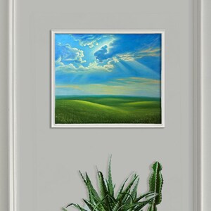 May include: A framed landscape painting depicts a vibrant green field under a bright blue sky with fluffy white clouds. The sun's rays pierce through the clouds, illuminating the scene. The artwork is mounted on a gray wall.
