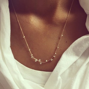 May include: A delicate gold necklace with a curved bar design and small round beads. The necklace is worn by a person with a white shirt.