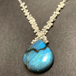 以下が含まれることがあります： 銀色のネックレス。洋梨型の、虹色の青いラブラドライトのペンダントが特徴です。ネックレスチェーンは、小さな白いビーズのクラスターで構成されています。ペンダントは、黒い静脈を持つさまざまな青の色合いを表示します。
