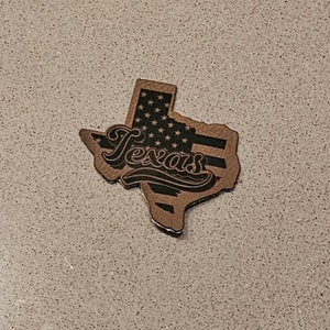 May include: A brown outline of the state of Texas with a black and white American flag design and the word "Texas" in a stylized font.