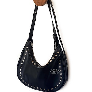 Puede incluir: Un bolso de mano de cuero negro con tachuelas plateadas colgado de un gancho de madera. El bolso tiene una forma curva y una cremallera. El nombre de la marca "AORA SABINE" est&aacute; impreso en la parte delantera del bolso.