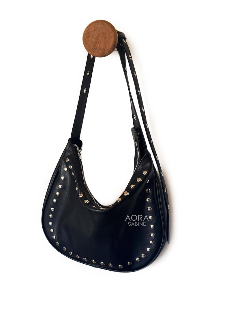 Puede incluir: Un bolso de mano de cuero negro con tachuelas plateadas colgado de un gancho de madera. El bolso tiene una forma curva y una cremallera. El nombre de la marca "AORA SABINE" est&aacute; impreso en la parte delantera del bolso.