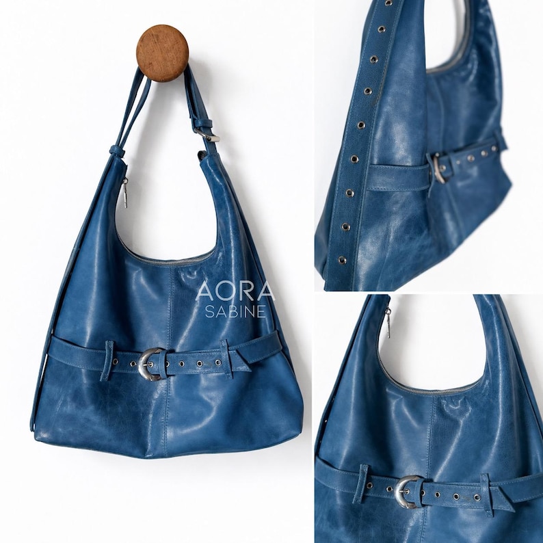 May include: A blue leather hobo bag featuring a horizontal strap with a silver-toned metal buckle across the front. The bag has a single shoulder strap and silver grommets along the side panel. The text AORA SABINE is printed in white.