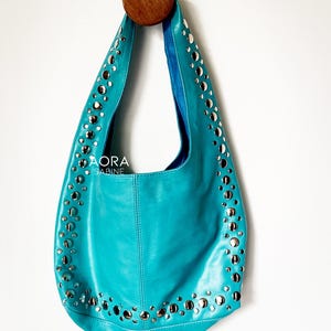 Puede incluir: Un bolso de mano de cuero azul turquesa con tachuelas plateadas alrededor de los bordes. El bolso tiene una sola correa y el nombre de la marca "AORA SABINE" está impreso en la parte delantera.