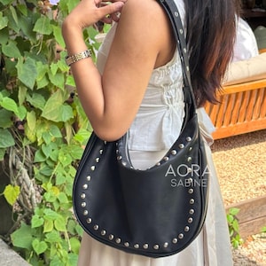 Puede incluir: Un bolso hobo de cuero negro con tachuelas plateadas y una correa negra ajustable. El bolso tiene el texto "AORA SABINE" en la parte delantera.