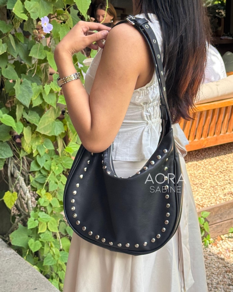 Puede incluir: Un bolso hobo de cuero negro con tachuelas plateadas y una correa negra ajustable. El bolso tiene el texto "AORA SABINE" en la parte delantera.