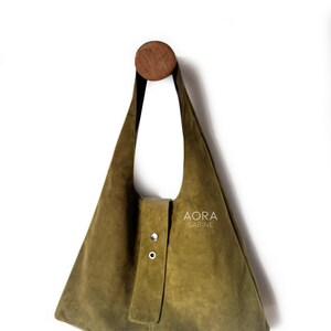 May include: Olive green suede shoulder bag with a triangular shape, hanging from a wooden peg. The bag features a flap closure with two silver-tone rivets and the text "AORA SABINE".