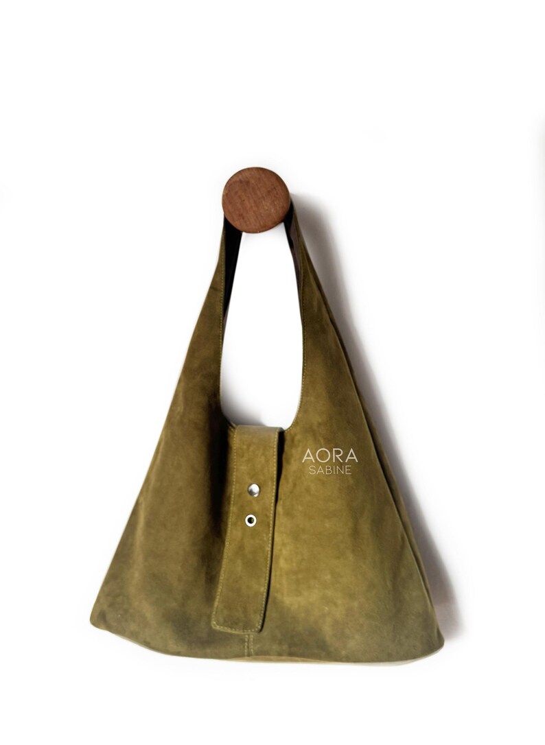 May include: Olive green suede shoulder bag with a triangular shape, hanging from a wooden peg. The bag features a flap closure with two silver-tone rivets and the text "AORA SABINE".