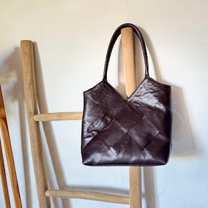 Può includere: Una borsa tote in pelle marrone scuro con un design intrecciato a forma di diamante. La borsa ha due manici lunghi e una parte superiore a forma di V. La borsa è appesa a un supporto di legno.