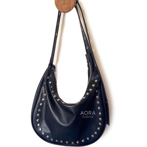 Puede incluir: Un bolso de mano de cuero negro con tachuelas plateadas colgado de un gancho de madera. El bolso tiene una forma curva y un cierre de cremallera. El nombre de la marca "AORA SABINE" est&aacute; impreso en la parte delantera del bolso.