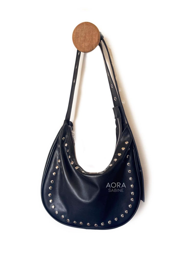 Puede incluir: Un bolso de mano de cuero negro con tachuelas plateadas colgado de un gancho de madera. El bolso tiene una forma curva y un cierre de cremallera. El nombre de la marca "AORA SABINE" est&aacute; impreso en la parte delantera del bolso.