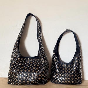 Puede incluir: Dos bolsos de hombro negros estilo hobo de diferentes tamaños, ambos con un patrón denso de tachuelas metálicas circulares de tono plateado que cubren toda la superficie exterior, incluidas las correas, con un acabado de cuero negro liso.