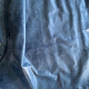 May include: A large, irregular piece of blue leather with a smooth, slightly matte finish and subtle tonal variations across its surface. The material features natural folds and creases, displaying a soft, flexible texture throughout the hide.