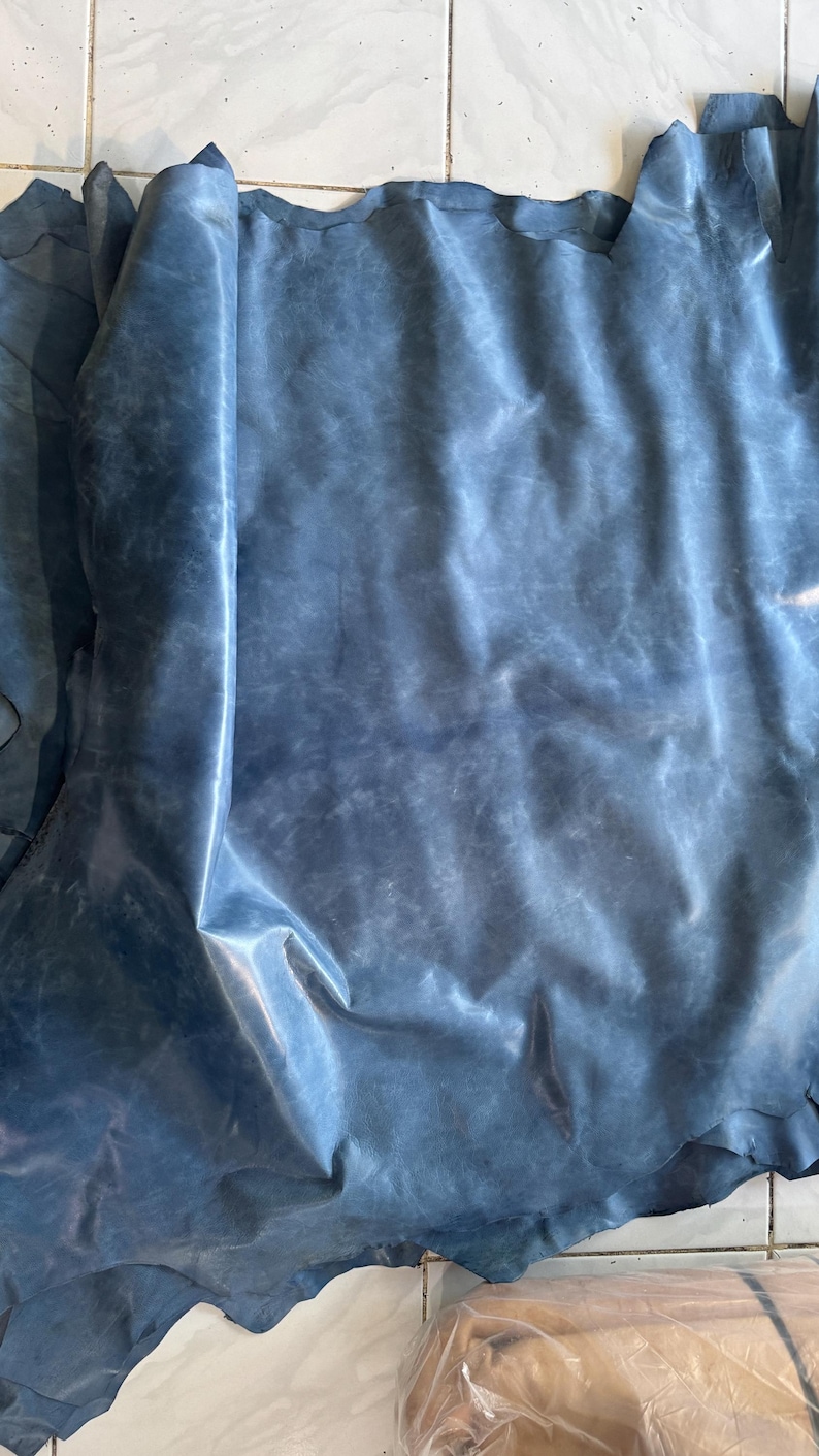 May include: A large, irregular piece of blue leather with a smooth, slightly matte finish and subtle tonal variations across its surface. The material features natural folds and creases, displaying a soft, flexible texture throughout the hide.