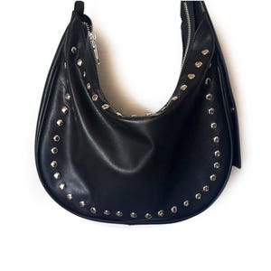 Puede incluir: Un bolso hobo de cuero negro con tachuelas plateadas alrededor de los bordes.