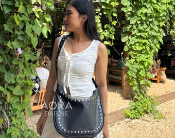 May include: A black leather shoulder bag with silver studs. The bag has a curved shape and a flap closure. The bag is worn by a person wearing a white top and a white skirt. The bag has the text "AORA SABINE" on the front.