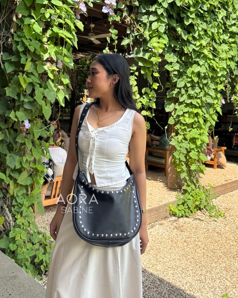 Puede incluir: Un bolso de hombro de cuero negro con tachuelas plateadas. El bolso tiene una forma curva y un cierre de solapa. El bolso lo lleva una persona que lleva una blusa blanca y una falda blanca. El bolso tiene el texto "AORA SABINE" en la parte delantera.