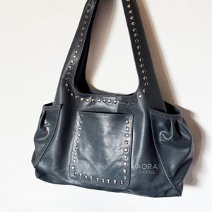 Puede incluir: Un bolso de hombro gris oscuro con tachuelas metálicas plateadas en la correa y el bolsillo frontal. El bolso incluye dos bolsillos laterales con detalles fruncidos y el texto AORA SABINE impreso en blanco en la parte inferior derecha.