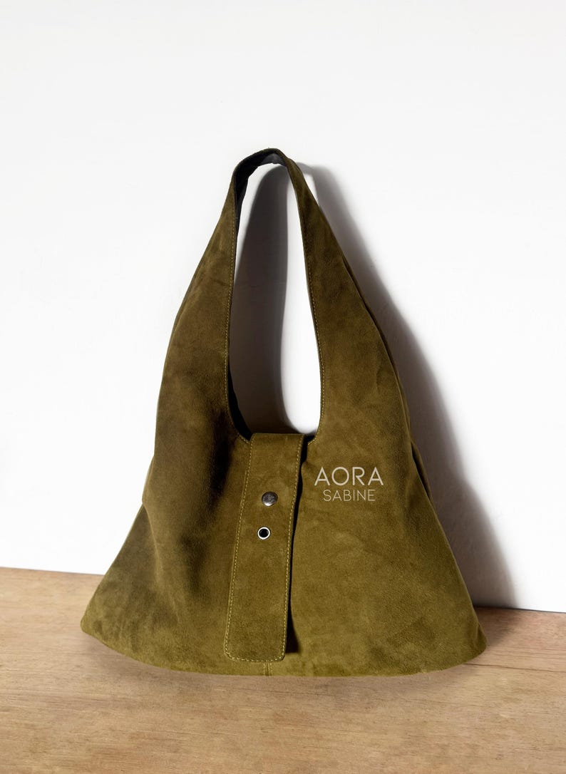 May include: Olive green suede shoulder bag with a unique, asymmetrical design. Features a single strap and a flap closure with two silver-tone rivets. The bag has the text "AORA SABINE" printed on the front.