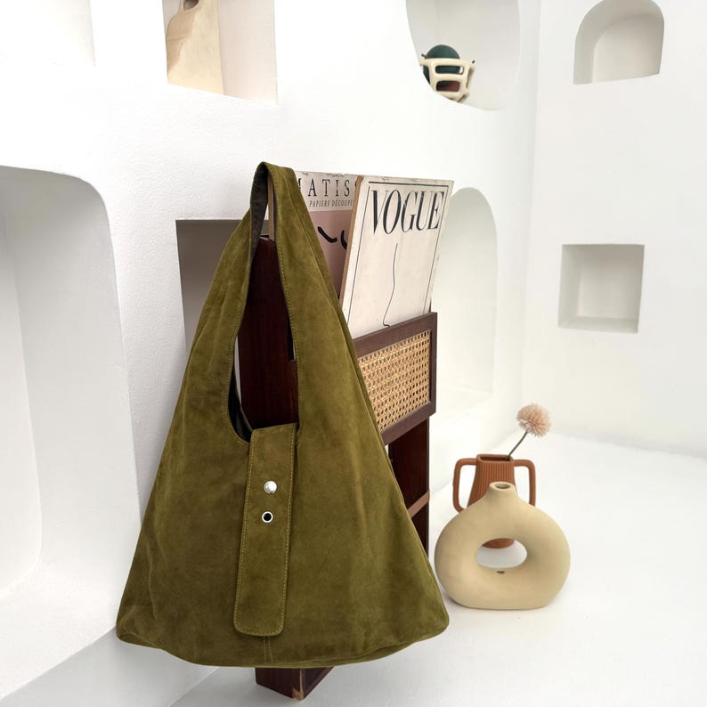May include: Olive green suede tote bag with a triangular shape and a single strap. The bag features a flap closure with two silver-tone snap buttons. The bag is hanging on a wooden rack next to a magazine holder.