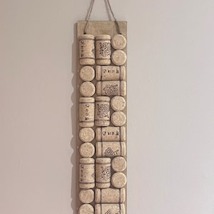 May include: A wooden board with a string for hanging, decorated with a vertical row of cork wine bottle stoppers. The stoppers have a grape design and the text "4121 B" printed on them.