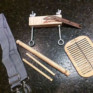 May include: A wooden loom kit with a gray strap, three wooden dowels, a wooden weaving needle, and a wooden loom with the text "HANDYMAN" printed on it.