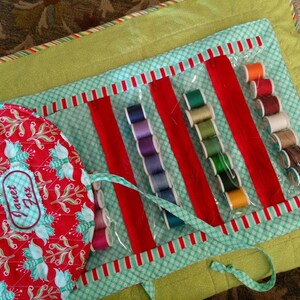 May include: A green and red fabric sewing roll with clear plastic pockets to hold spools of thread. The roll has a red fabric flap with the words "Project Box" embroidered on it.