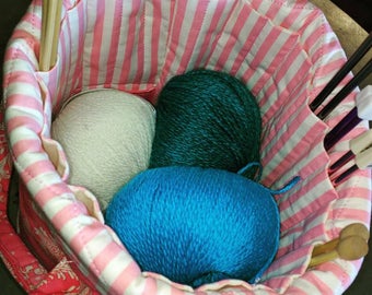 May include: A pink and white striped fabric tote bag with three balls of yarn inside. The yarn is teal, white, and blue. There are also wooden knitting needles in the bag.