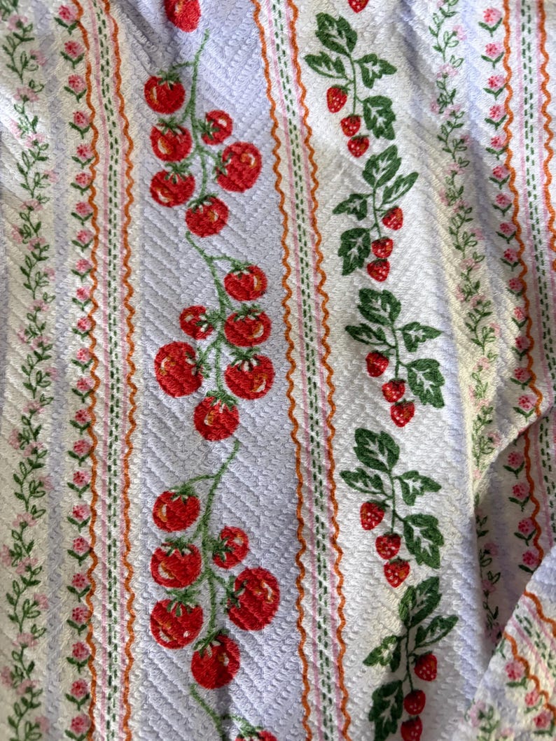 May include: Textile featuring a repeating pattern of red tomatoes and green leaves on a pale blue backdrop. Vertical stripes with pink flowers and orange lines border the tomato and strawberry designs.
