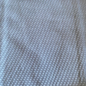 May include: Close-up of a pale grey woven fabric with a textured, honeycomb-like pattern. The fabric appears soft and pliable, with subtle variations in tone across its surface. The weave creates a grid of small, raised squares.