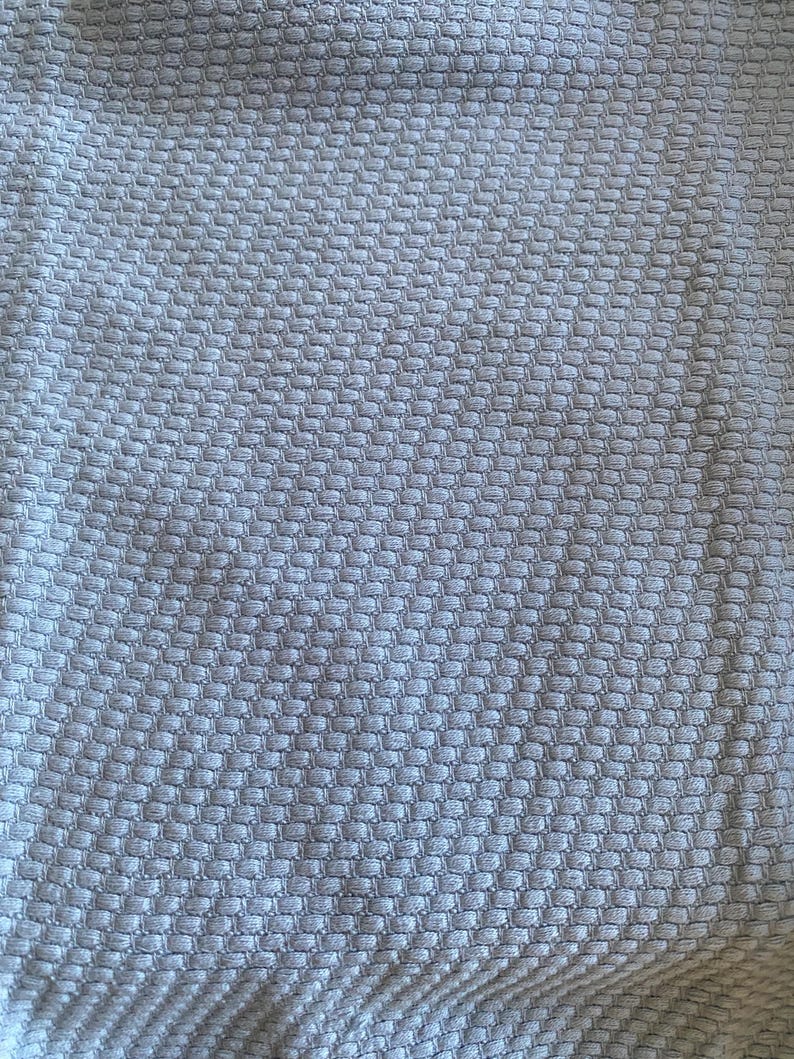 May include: Close-up of a pale grey woven fabric with a textured, honeycomb-like pattern. The fabric appears soft and pliable, with subtle variations in tone across its surface. The weave creates a grid of small, raised squares.