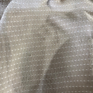 May include: Close-up of a beige fabric with a dotted pattern. The fabric has a woven texture, and the white dots are evenly spaced across the surface. The fabric appears to be soft and pliable, with slight folds and wrinkles.