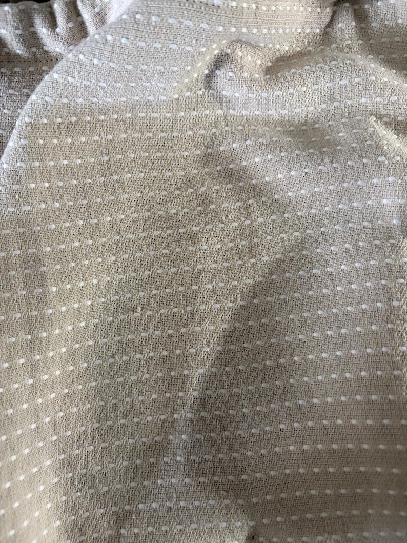 May include: Close-up of a beige fabric with a dotted pattern. The fabric has a woven texture, and the white dots are evenly spaced across the surface. The fabric appears to be soft and pliable, with slight folds and wrinkles.